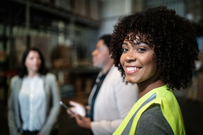 A smiling woman with curly hair wears a bright yellow safety vest in a warehouse setting. Two other women are blurred in the background, engaged in conversation. The focus is on the woman in the foreground, who exudes confidence and friendliness.