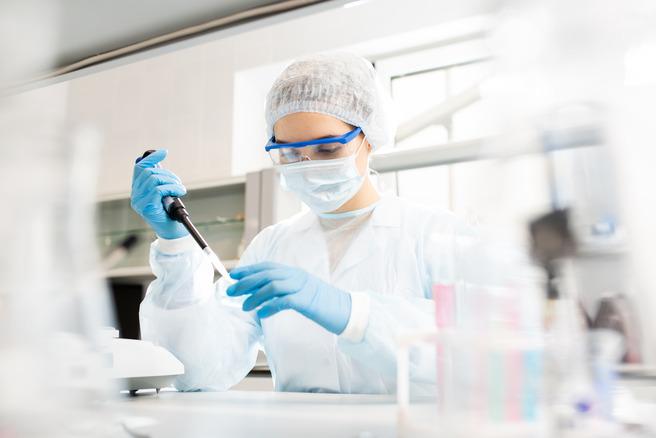 A laboratory technician in protective gear, including gloves, a lab coat, a face mask, and goggles, is using a pipette to handle a liquid sample on a lab bench surrounded by laboratory equipment.