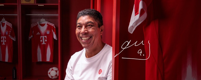 A smiling man sits in a red locker room, surrounded by Bayern Munich jerseys. The image conveys a sense of joy and pride in a football environment. A signature is displayed in the corner, adding a personal touch.