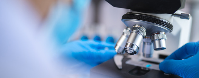 A close-up of a laboratory microscope with multiple objective lenses, as a person wearing blue gloves adjusts the microscope. The background is slightly blurred, suggesting an active lab environment.