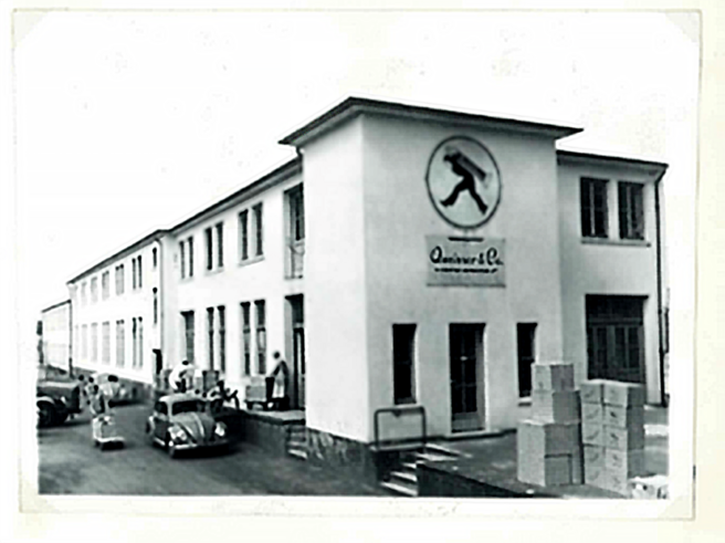 A historical black-and-white photo of a two-story building featuring a logo of a person in motion. Boxes are stacked on the sidewalk, and a few people are seen near parked cars. The building appears to be a factory or warehouse, with multiple windows and a sign above the entrance.