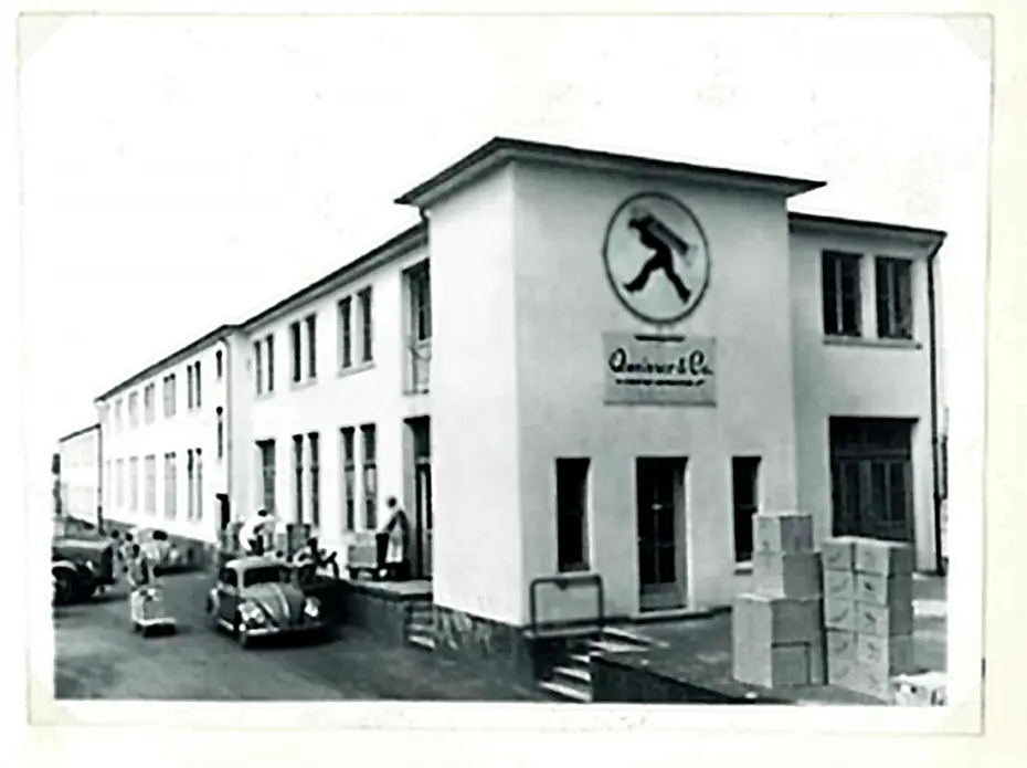 A historical black-and-white photo of a two-story building featuring a logo of a person in motion. Boxes are stacked on the sidewalk, and a few people are seen near parked cars. The building appears to be a factory or warehouse, with multiple windows and a sign above the entrance.