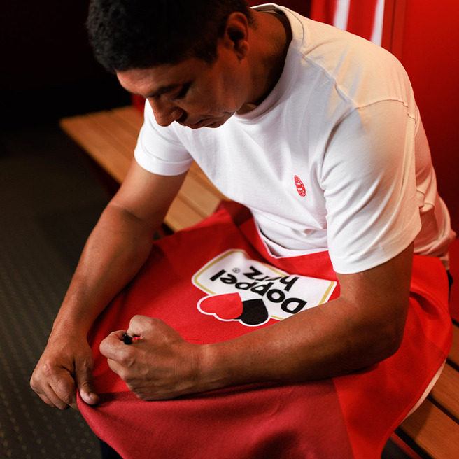 A person is sitting on a wooden bench in a locker room, focused on tying the laces of a red sports garment with a logo that says "Popped Pix." They are wearing a white t-shirt and appear to be preparing for an activity.