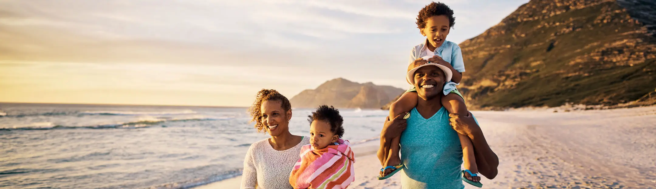 A family stands on a beach at sunset. Two adults are smiling, one holding a child, while another child is on an adult's shoulders. The ocean and mountains are visible in the background, creating a warm and joyful atmosphere.