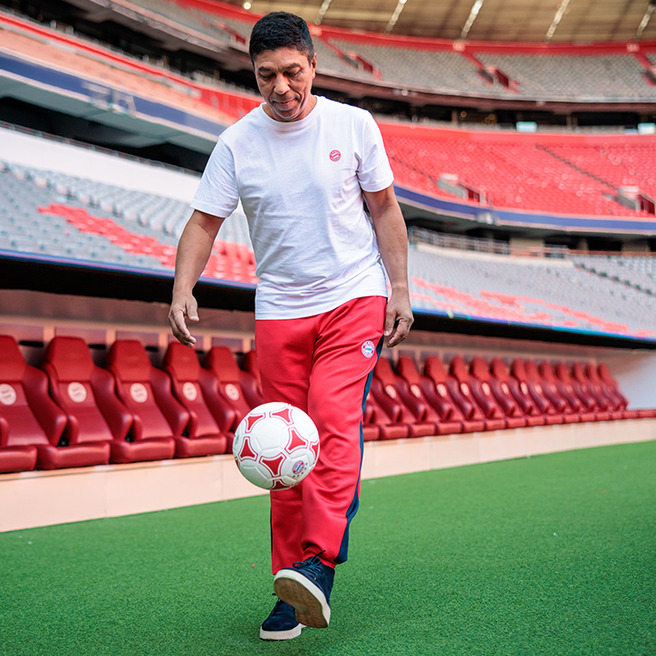 A man in a white t-shirt and red athletic pants skillfully juggles a soccer ball on the field of a stadium. Red seating is visible in the background, indicating a sports venue setting.