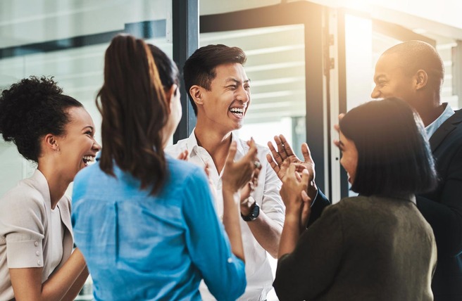 A group of five diverse individuals is joyfully engaged in conversation, laughing and applauding each other in a bright, modern indoor setting. The atmosphere is cheerful and collaborative, reflecting camaraderie and positivity.
