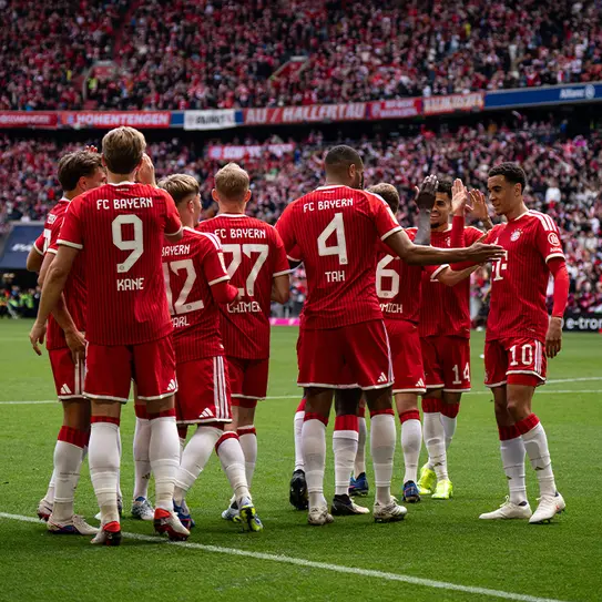 A group of FC Bayern Munich football players in red uniforms gather together on the field, some high-fiving and celebrating. The backdrop shows a packed stadium with fans. The players display teamwork and enthusiasm during what appears to be a match.