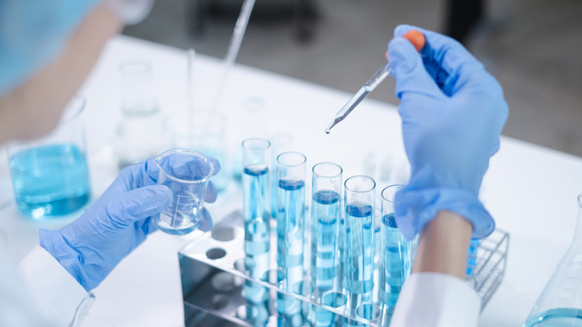 A laboratory scene shows a researcher in gloves using a pipette to transfer a blue liquid into test tubes. A small beaker containing liquid is held in the other hand, with more glassware in the background.