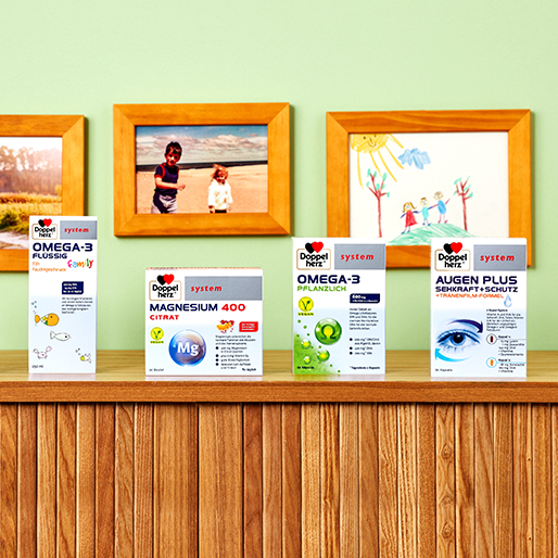 Five boxes of dietary supplements are displayed on a wooden shelf against a colorful wall. The products include Omega-3 liquid, Magnesium 400 Citrate, and Omega-3 plant-based, alongside a vision supplement labeled Augen Plus. Family photos and children's drawings decorate the wall above the shelf.