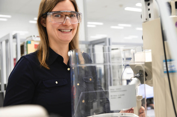 A smiling woman wearing safety glasses stands beside laboratory equipment. She is dressed in a dark shirt and appears engaged in her work environment, which features a bright, modern laboratory setting.