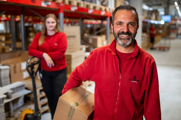 A smiling man in a red jacket holds a box in a warehouse, while a woman in a similar red jacket stands in the background beside a pallet jack. The space is filled with stacked boxes and shelves, indicating a busy work environment.