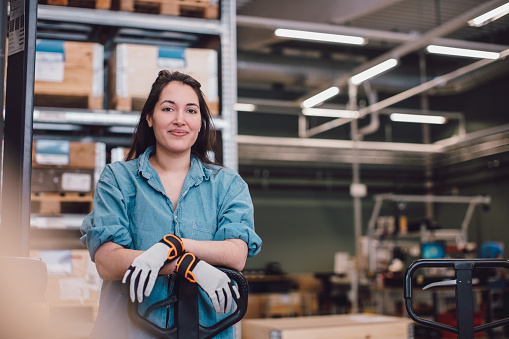 A woman in a blue button-up shirt and gloves stands confidently with her arms crossed, smiling. She is positioned in a warehouse setting, with shelves filled with wooden boxes in the background, suggesting a work environment.